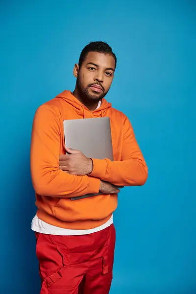 Чарівний афроамериканський чоловік у яскравому вбранні тримає руки з ноутбуком на синьому фоні — стокове фото
