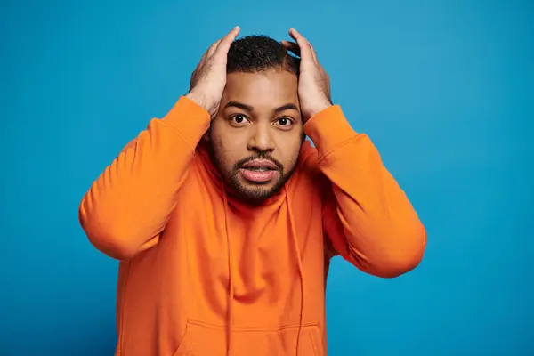 Worried african american young man in orange outfit holding to head with hands on blue background — Stock Photo