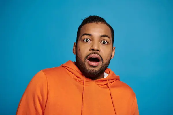 Atractivo hombre afroamericano en traje naranja se sorprendió contra fondo azul - foto de stock