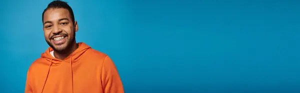 Estandarte de hombre americano africano alegre en traje naranja sonriendo ampliamente sobre fondo azul - foto de stock