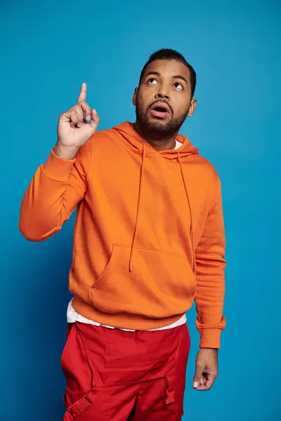 Nachdenklicher afrikanisch-amerikanischer Mann in orangefarbenem Outfit, der vor blauem Hintergrund den Finger nach oben reckt — Stockfoto