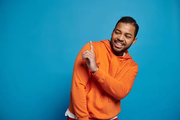 Smiling african american man in vibrant outfit putting finger and looking to side on blue background — Stock Photo