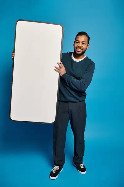 Atractivo afroamericano joven sosteniendo maqueta de teléfono inteligente en las manos contra el fondo azul - foto de stock