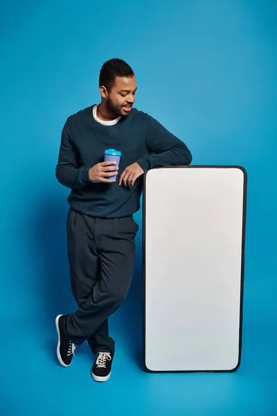 Jeune homme afro-américain détendu tenant une tasse en papier et se reposant près de la bannière de maquette de smartphone — Photo de stock