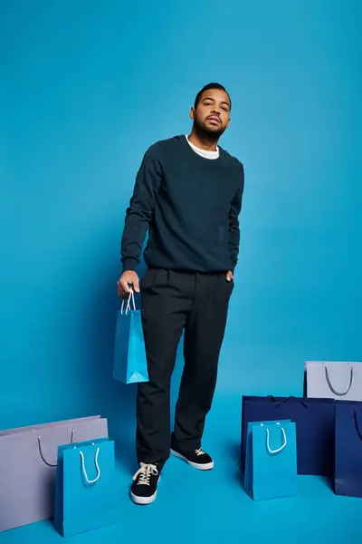 Encantador hombre afroamericano en traje azul oscuro de pie con bolsas de compras y mano en el bolsillo - foto de stock