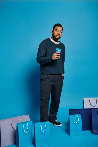Hombre afroamericano en traje azul oscuro de pie con bolsas de compras, taza de papel y mano en el bolsillo - foto de stock