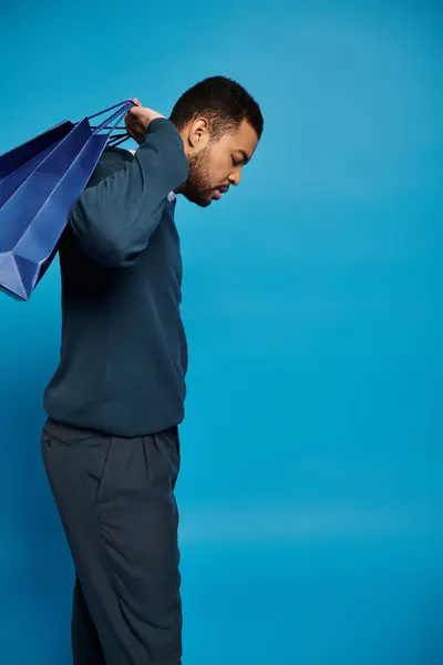 Homme afro-américain en tenue bleu foncé regardant vers le bas et tenant des sacs à provisions au-dessus de l'épaule — Photo de stock