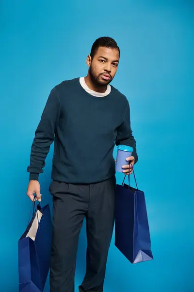 Encantador hombre afroamericano sosteniendo bolsas de compras con taza de papel en la mano sobre un fondo vibrante - foto de stock