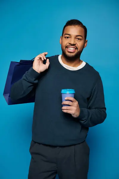 Улыбающийся африканский американец держит бумажную чашку с пакетом для покупок через плечо на синем фоне — стоковое фото