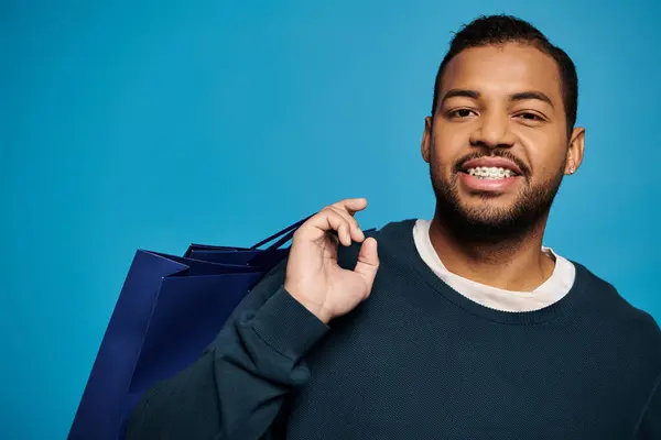 Retrato de joven afroamericano alegre con bolsa de compras sobre el hombro sobre fondo azul - foto de stock
