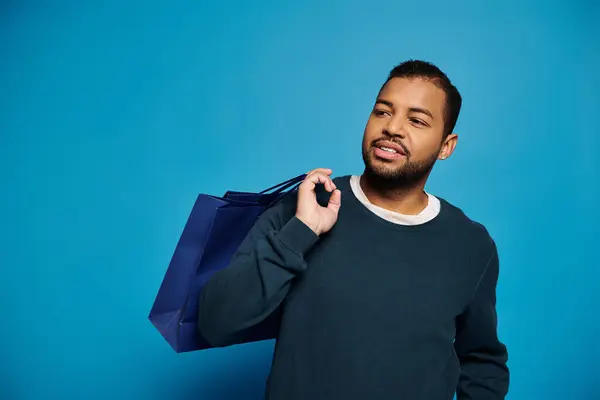 Charismatische afrikanisch-amerikanische Mann mit Blick auf die Seite Einkaufstasche über der Schulter auf blauem Hintergrund — Stockfoto