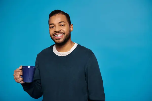 Homme afro-américain joyeux dans sa vingtaine tenant la tasse à la main sur fond bleu — Photo de stock