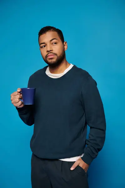 Харизматичный африканский американец в возрасте 20 лет держит чашку в руке с рукой в кармане на синем фоне — стоковое фото