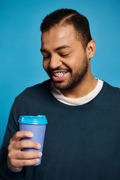 Retrato del joven afroamericano sonriente mirando a la taza de papel en la mano sobre fondo azul - foto de stock