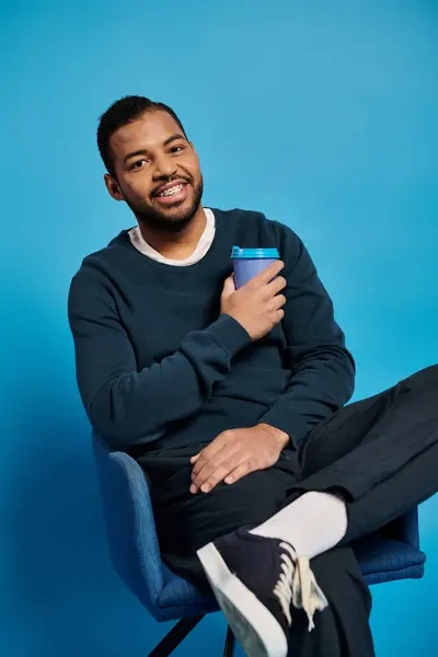 Atractivo afroamericano hombre en 20s sentado en silla con taza de papel sobre fondo azul - foto de stock