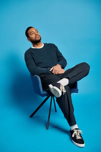 Handsome african american man in his 20s sitting on chair with folded arms on blue background — Stock Photo