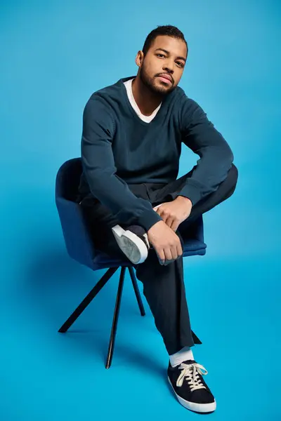 Encantador homem afro-americano em seus 20 anos sentado na cadeira e inclinado para dobrar a perna no fundo azul — Fotografia de Stock