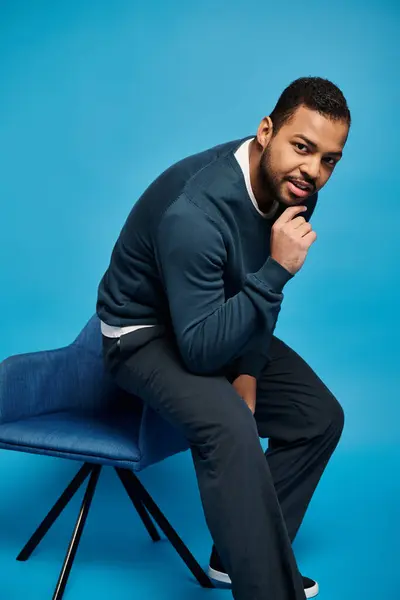 Encantador hombre afroamericano sentado en el respaldo de la silla y tocando la barbilla sobre fondo azul - foto de stock
