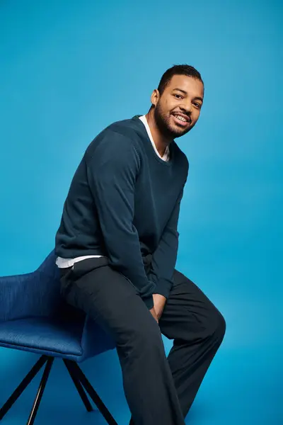 Handsome african american man in dark outfit sitting on back of chair against blue background — Photo de stock