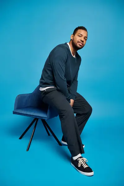 Atractivo afroamericano hombre en traje oscuro relajado sentado en el respaldo de la silla sobre fondo azul - foto de stock