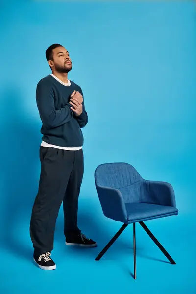 Очаровательный африканский американец с руками на груди, стоящими рядом со стулом на синем фоне — стоковое фото