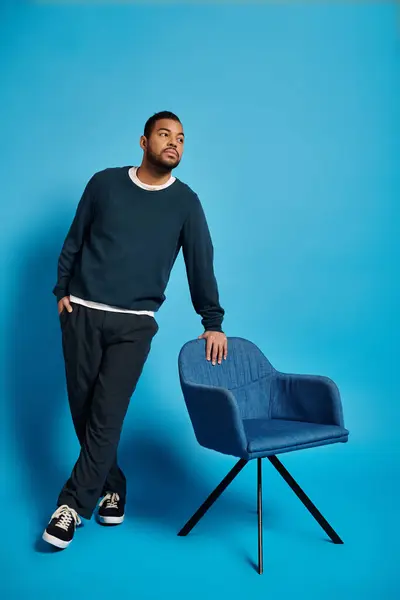 Guapo afroamericano hombre inclinado a silla y mirando a un lado sobre fondo azul - foto de stock