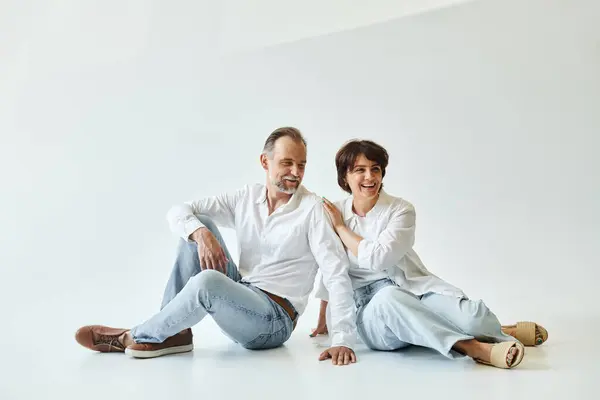 Middle aged couple wearing white shirts and jeans sitting on grey background and looking away — Stock Photo