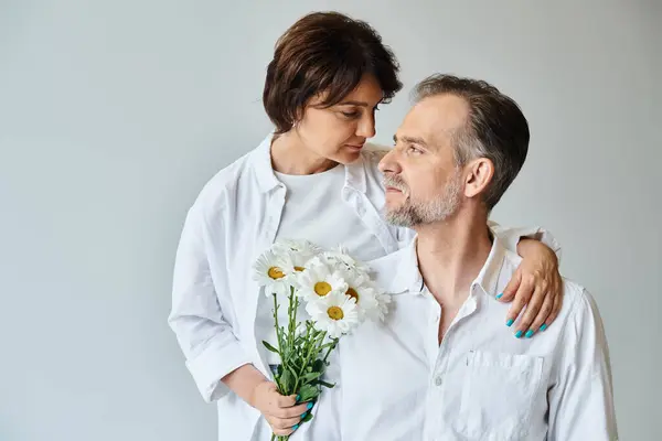 Portrait de couple mature heureux avec des fleurs sur fond gris, femme étreignant et s'appuyant sur l'homme — Photo de stock