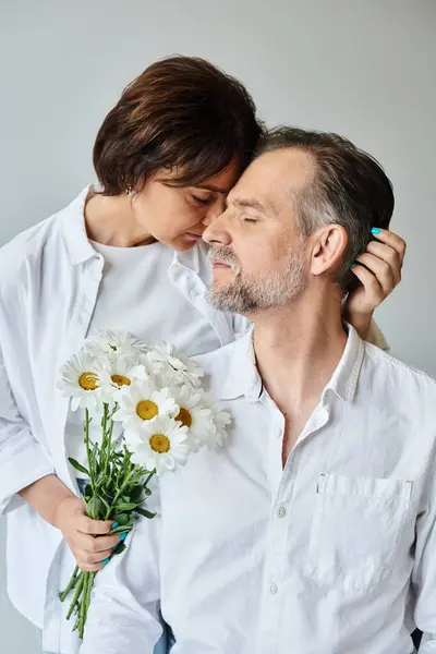 Mulher atraente segurando flores e abraçando homem maduro em camisa branca no fundo cinza — Fotografia de Stock