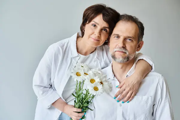 Porträt eines Paares mittleren Alters mit Blumen auf grauem Hintergrund, Frau umarmt und lehnt sich an Mann — Stockfoto