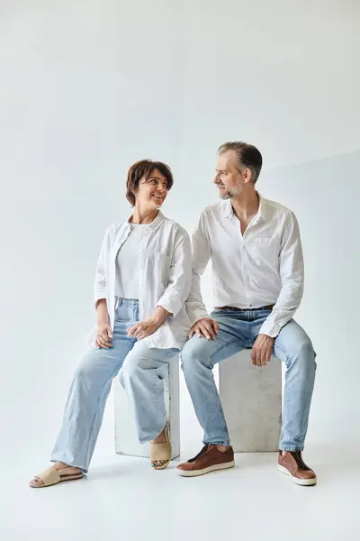 Ganzes Porträt eines Paares mittleren Alters, das auf grauem Hintergrund sitzt und einander ansieht — Stockfoto