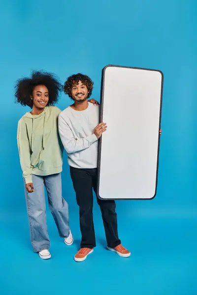 Ein bunt gemischtes Studentenpaar steht Seite an Seite neben einer Smartphone-Attrappe in einem Atelier und präsentiert kulturelle Vielfalt auf blauem Hintergrund. — Stockfoto