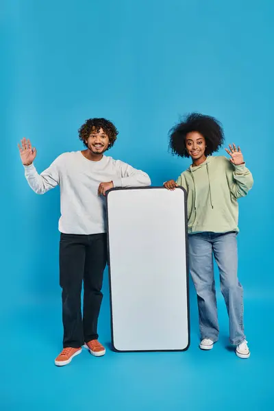 Dois estudantes multiculturais de pé ao lado um do outro, segurando um sinal em um estúdio com um fundo azul, mostrando diversidade cultural. — Fotografia de Stock