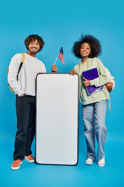 Um par de indivíduos diversos, estudantes multiculturais, estão perto de um mockup smartphone em um estúdio contra um fundo azul. — Fotografia de Stock
