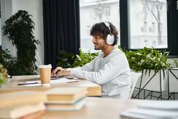 Indischer Mann, der an einem Tisch sitzt, in seinen Laptop vertieft, Kopfhörer trägt und in seine Arbeit in einem modernen Coworking Space eintaucht. — Stockfoto