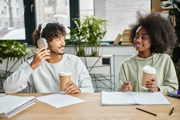 A man and woman of different cultures sitting at a table, sipping coffee and engaging in conversation in a modern coworking space. — Stock Photo