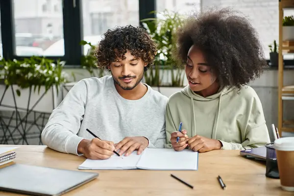 A man and a woman of different ethnic backgrounds sit at a table, writing together in a modern coworking space. — Stock Photo