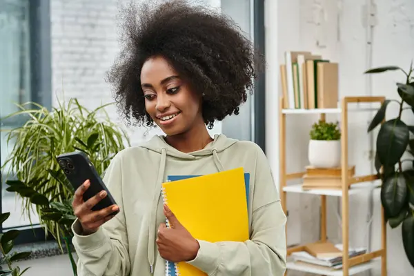 Una mujer de varias etnias sostiene una carpeta mientras envía mensajes de texto en un teléfono inteligente en un espacio de coworking moderno. - foto de stock
