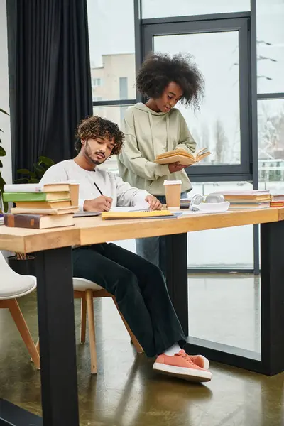 Dos estudiantes de diferentes orígenes culturales se sientan en una mesa, absortos en libros y notas en un moderno espacio de coworking. - foto de stock