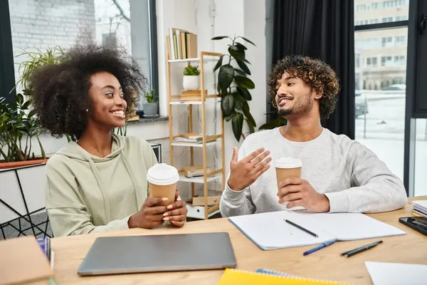 Un hombre y una mujer de diferentes culturas conversan mientras están sentados en una mesa en un moderno espacio de coworking. - foto de stock