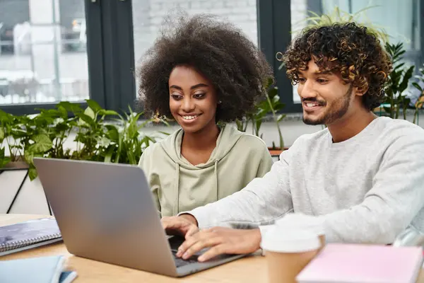 A man and woman of different cultural backgrounds work side by side at a table with a laptop in a modern coworking space. — Stock Photo