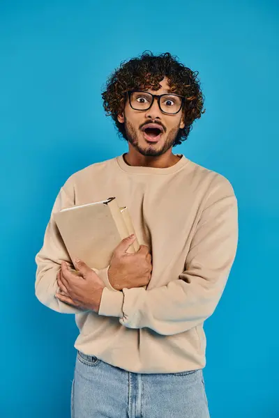 Studente indiano scioccato con capelli ricci e occhiali, in piedi in abbigliamento casual, tenendo una cartella su uno sfondo blu in uno studio. — Foto stock