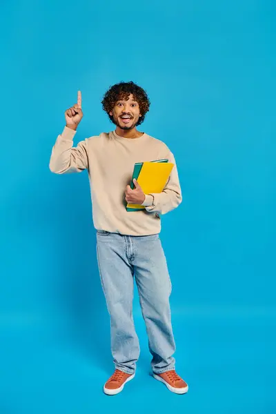 Um estudante indiano em traje casual está contra um pano de fundo azul, segurando um livro em uma mão e fazendo um sinal de paz com a outra.. — Fotografia de Stock