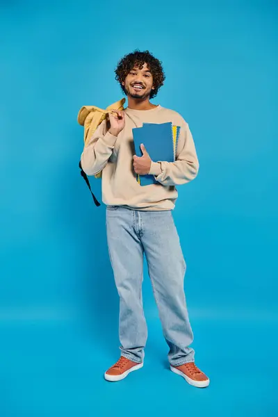 A young Indian student holding a backpack and a book in a casual attire against a blue backdrop. — Stock Photo