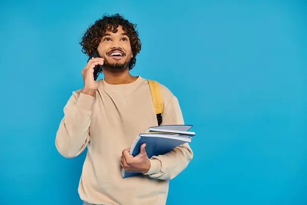 Un estudiante indio con atuendo casual se levanta contra un telón de fondo azul, sosteniendo una carpeta y hablando en un teléfono celular. - foto de stock