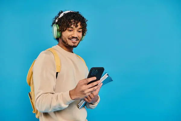An Indian student standing, wearing headphones, and holding a book against a blue backdrop in a studio setting. — Stock Photo