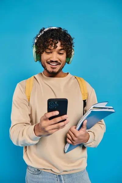 Uno studente indiano in abbigliamento casual ascolta musica con le cuffie mentre tiene un telefono cellulare su uno sfondo blu. — Foto stock