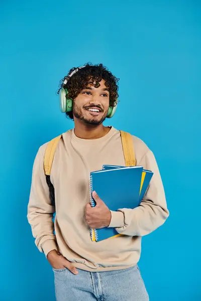 Un estudiante indio está parado sobre un telón de fondo azul, usando auriculares y sosteniendo un libro, una mezcla armoniosa de música y literatura. - foto de stock