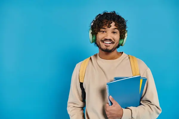 Happy indian man wearing headphones, holding books and smiling on blue background — Stock Photo