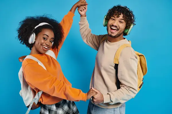 An interracial man and woman dance together, wearing headphones, expressing joy and connection through movement on a blue backdrop. — Stock Photo